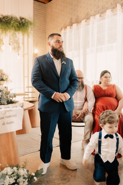 This image shows a groom dressed in a fitted navy blue suit with white shoes, standing inside a well-lit venue with large windows and sheer white curtains. The groom has a full beard and a short hairc