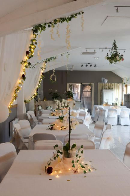 A beautifully decorated indoor venue featuring soft, ambient lighting with fairy lights and hanging floral arrangements. The space includes tables draped in white tablecloths, each adorned with elegan