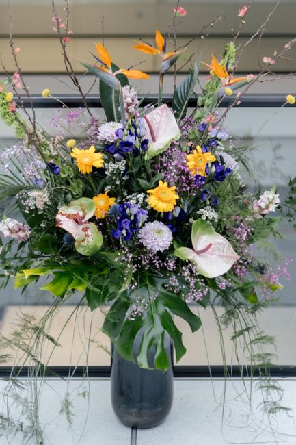 A large, vibrant floral arrangement in a tall, dark glass vase situated on a window ledge, featuring a variety of fresh flowers including yellow daisies, white calla lilies with pink accents, purple c