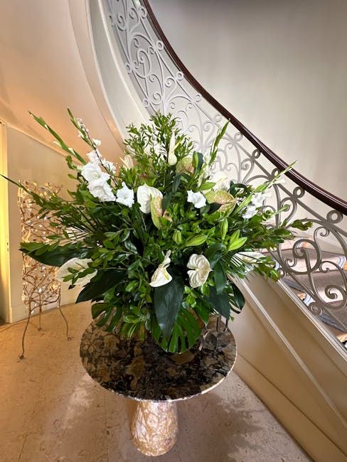 A large floral arrangement featuring white lilies, anthuriums, and various green foliage, placed in a dark ceramic vase atop a marble-topped table near a staircase with ornate white balustrades. The b