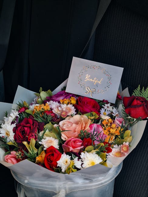 A vibrant bouquet of fresh flowers arranged in a round, white paper wrapping, featuring red roses, pink carnations, white daisies, yellow solidago, and purple statice. The bouquet is positioned on a c