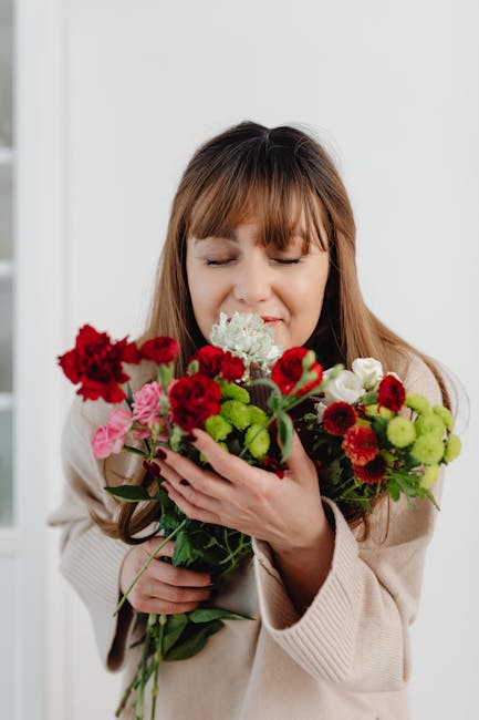 A woman with light brown hair and bangs gently holds a vibrant floral bouquet in front of her face, eyes closed with a content expression. The bouquet features a variety of fresh flowers including red